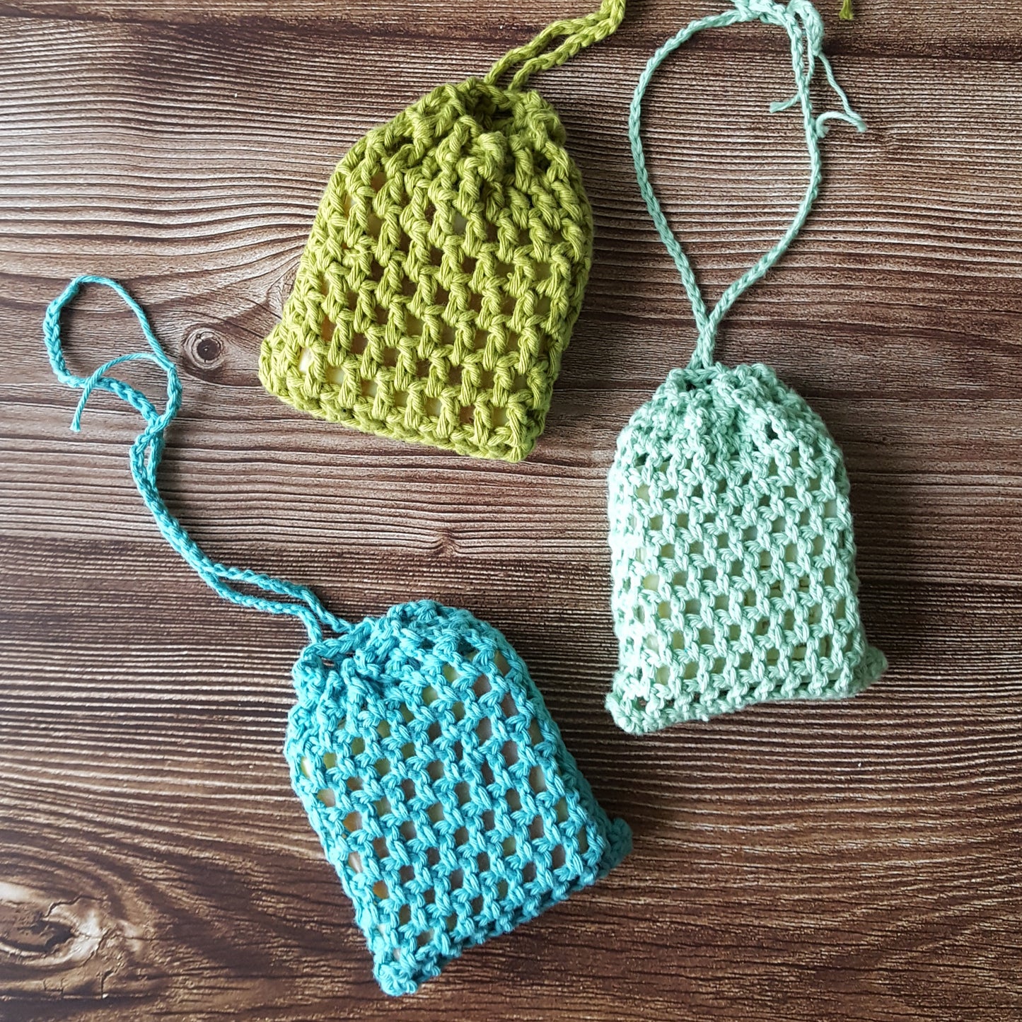 Trois pochettes pour savon de couleur vert olive, menthe et turquoise sont présentés sur une table en bois. Les pochettes de savon permettent de garder votre savon sec entre deux utilisations. Crocheté à la main en France à partir de coton biologique par ILELI savons, Orsay, granny style, tout doux, salle de bain zero dechet